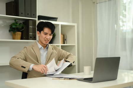 Frustrated businessman holding paperwork sitting at desk with laptop.の写真素材