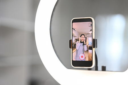 Close up of a smartphone mounted on a stand with ring light, capturing a woman recording a video in...