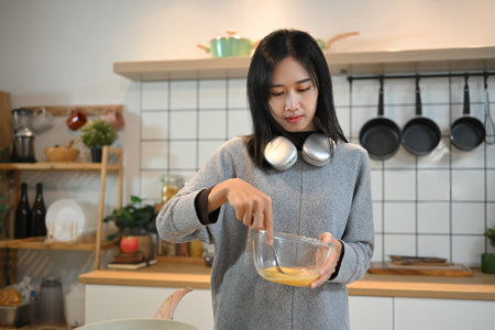 Young asian woman beating eggs in a bow with fork, making breakfast in the kitchen.