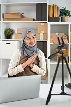 Young Muslim woman sitting in front of a smartphone mounted on a tripod, recording video or live streaming for her audience.の写真素材