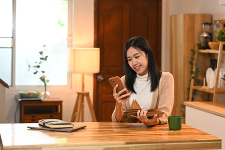 Beautiful young woman with book in hand using mobile phone at kitchen table.の写真素材