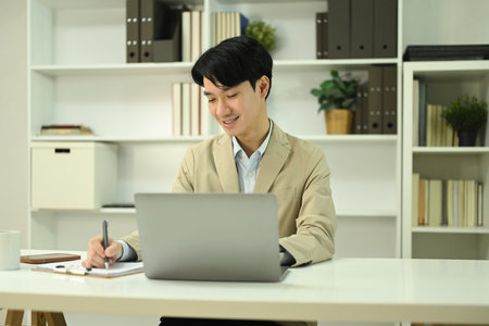 Young businessman in trendy suit using laptop and writing notes at office desk.の写真素材