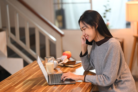 Smiling young female freelancer talking on mobile phone and using laptop at kitchen table.の写真素材