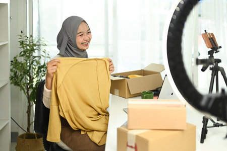 Young woman in a hijab conducting a live streaming showcasing a product to her audience. E-commerce and online retail concept.の写真素材