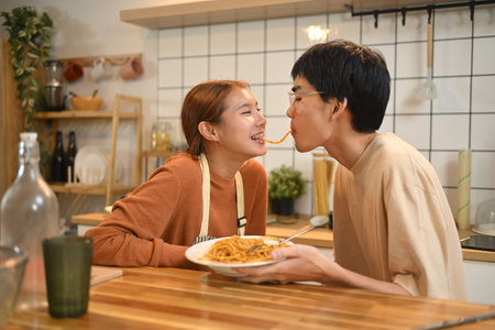 Romantic asian couple enjoying a meal, sharing spaghetti during dinner in cozy kitchen.の写真素材