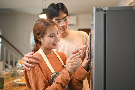 Happy asian couple interacting with digital tablet mounted on the door of a refrigerator.の写真素材