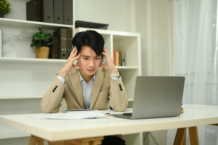 Stressed businessman holding head in his hands in front of laptop and financial reports.の写真素材