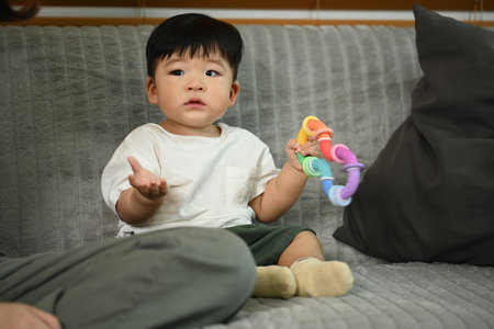 Cute little toddler boy sitting on comfortable couch, holding a colorful plastic toy in hand....