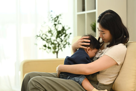 Young mother sitting on sofa, gently cradling her baby in her arms. Parenting, child care and family concept.の写真素材