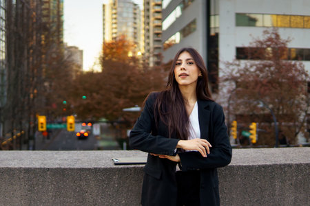 Confident hispanic businesswoman standing in front of a modern building with autumn foliage.の写真素材