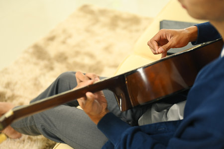 Closeup man is playing a guitar on a couch. He is wearing a blue shirt and gray pantsの写真素材