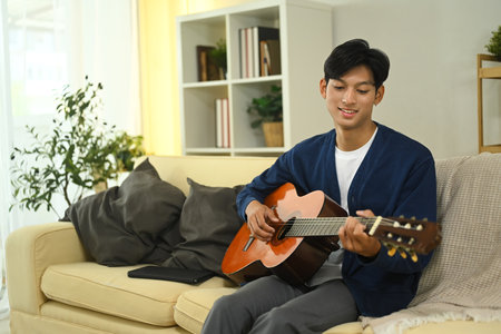 Smiling young adult man tuning and practicing acoustic guitar on sofa.の写真素材