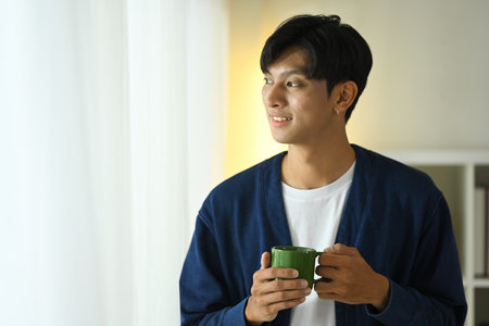Relaxed young adult man holding a coffee cup gazing outside with a smile.の写真素材