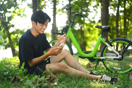 Smiling young man using mobile phone resting on the grass near his bike in a tranquil park.