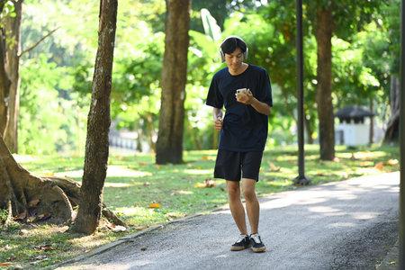 Young man holding mobile phone jogging along a paved path in a park on a sunny day.の写真素材
