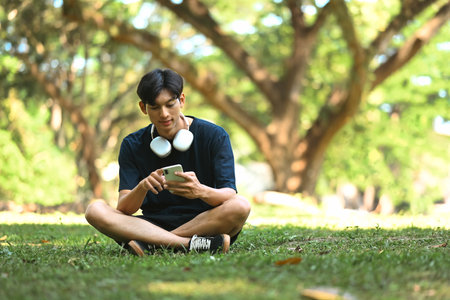 Young man sitting on the grass and using mobile phone in a park with sunlight filtering through the...
