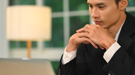 Serious and thoughtful businessman holding hands under chin looking at his laptop.の写真素材