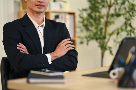 Confident businessman wearing a black suit sitting with his arms crossed in modern office.の写真素材