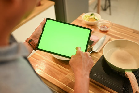 Close up of senior man using digital tablet in a cozy home kitchen to follow a recipe or cooking instructions.の写真素材