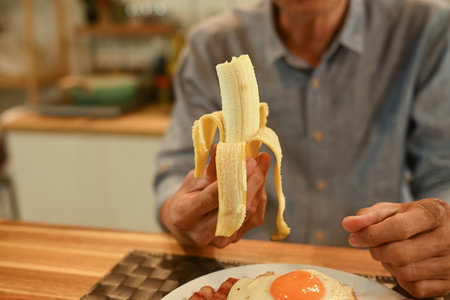 Senior man holding peeled banana while enjoying breakfast at kitchen table..の写真素材