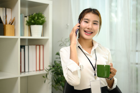 Pretty asian female employee holding cup of coffee while talking on smartphone in office..の写真素材