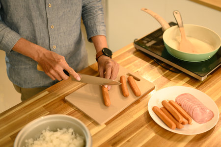 Cropped shot of man preparing simple meal in a kitchen, slicing sausages on a wooden cutting board.の写真素材