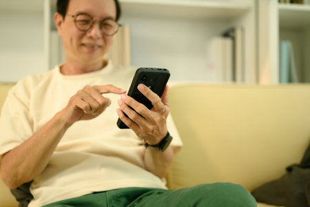 Happy elderly Asian man in eyeglasses using smartphone on couch at home.の写真素材