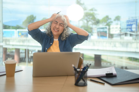 Stressed senior businesswoman holding her head in frustration while sitting at desk with laptop.の写真素材