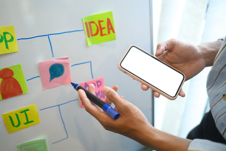 Designer holding smartphone with a blank screen mapping out app ideas on a whiteboard.