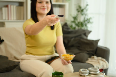Cheerful woman holding remote control and a bowl of chips watching tv in living room.の写真素材