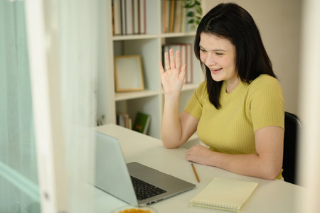 Smiling woman waving hand looking at laptop during virtual video conference call in home.