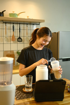 Cheerful young woman preparing a protein shake at wooden kitchen counter.の写真素材