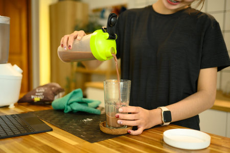 Smiling woman in sportswear pouring a freshly made chocolate protein shake into a glass.の写真素材