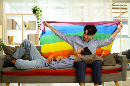 Cheerful gay couple holding a rainbow pride flag relaxing on sofa.の写真素材