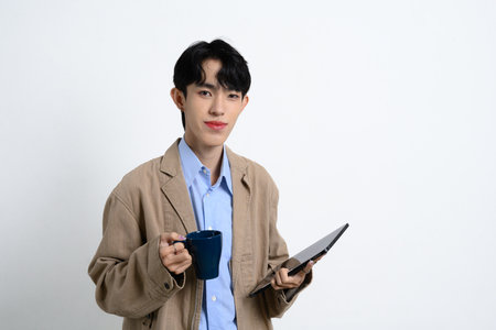 Confident young businessman holding a coffee mug and digital tablet standing over white background.の写真素材