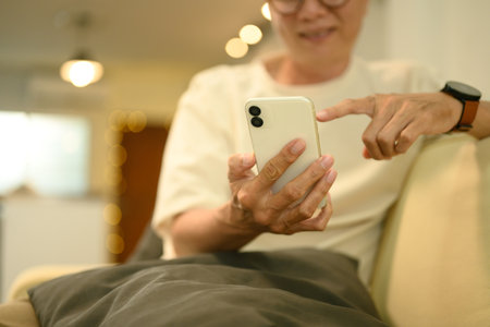 Happy elderly Asian man in eyeglasses using smartphone on sofa at home.の写真素材