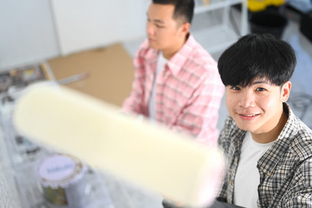 Two young asian men painting walls with a roller brush during renovation.