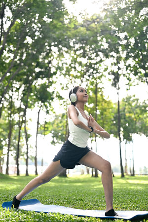 Serene African American woman wearing headphones practicing yoga on mat in a park.