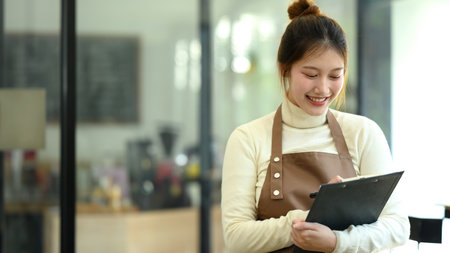 Smiling female waitress wearing an apron taking orders on a clipboard in cozy restaurant.の写真素材