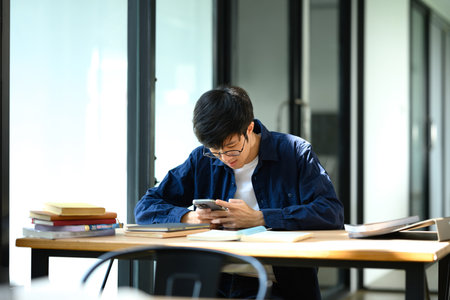 Student man using a smartphone at wooden desk surrounded by study materials.の写真素材