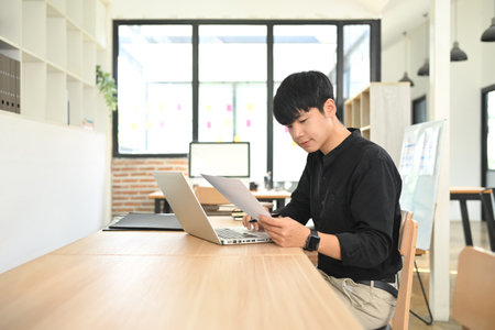 Person in a black shirt reviewing papers while working on a laptop in modern office.の写真素材