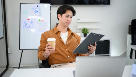 Handsome businessman holding a coffee cup and checking financial document at desk.