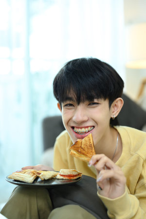 Cheerful young man enjoying homemade sandwiches in bright living room.
