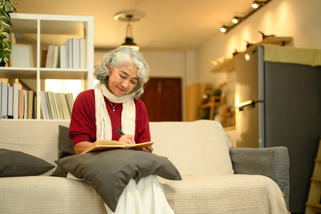 Calm senior woman in a red sweater sitting on couch and writing in journal.の写真素材