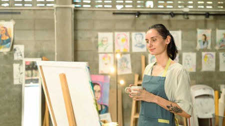 Female artist in an apron holding a cup of coffee standing in front of a canvas in a creative...