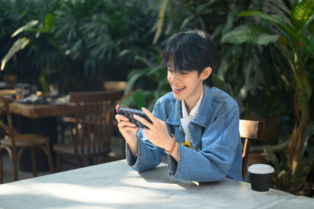 Young man sitting at a cafe table, smiling brightly while playing a game on smartphone.の写真素材