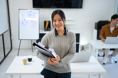 Portrait of smiling young woman standing in a bright office holding stack of documents and binders.の写真素材