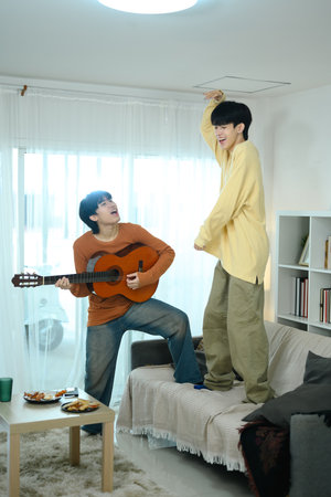 Cheerful gay couple enjoying casual moment at home playing the guitar and singing in living room.の写真素材
