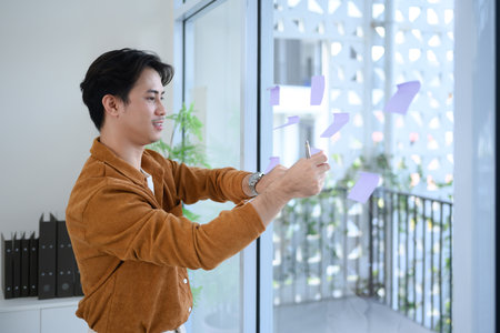 Young businessman sticking notes on a glass wall for project planning and organization.