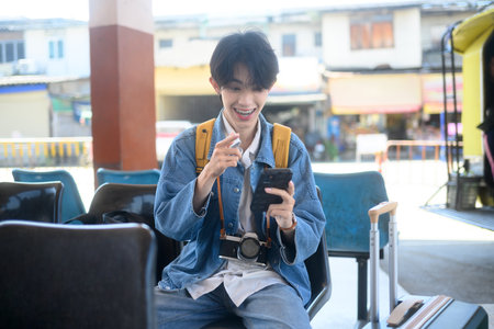 Young traveler with backpack checking travel plans on smartphone while sitting at transportation terminal.の写真素材
